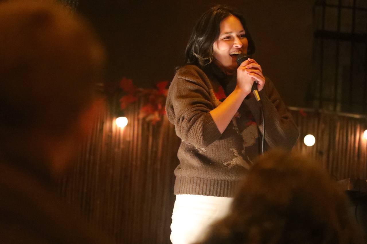 Willa doing standup at Warm Regards, mic in hand, under string lights